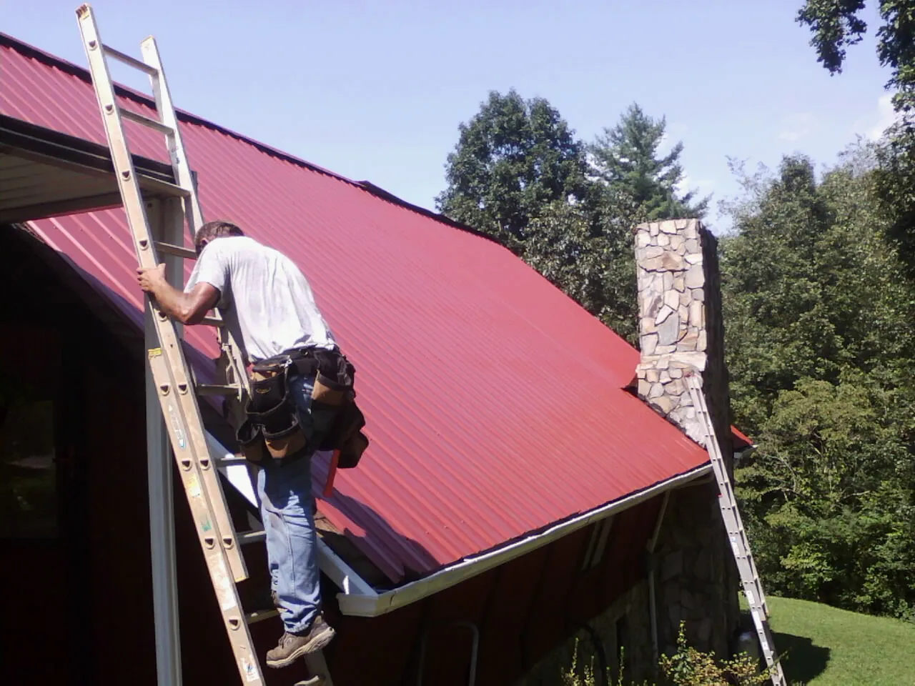Roofer on a ladder beside a red metal roof and stone chimney — Soffit & Fascia Repair by Roofing Brothers in Yorkville